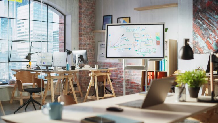 Modern office workspace with computers, a digital whiteboard displaying project planning diagrams, and collaborative workstations in a bright, open office.